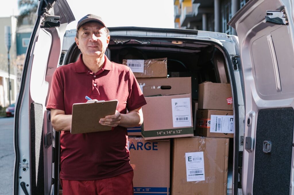 Elderly delivery person holds clipboard in front of open van filled with packages on a sunny day.